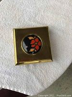 Top view of square brass box showing floral motif inset in circular glass-like medallion on lid.