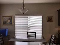 Photo showing dining area with two framed artworks hung on walls, one on each side of window blinds above a dining table and chairs.