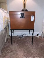 Front view of the wood storage box on black metal stand positioned beside a bed, showing the latch and overall condition of the wood surface.