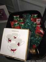 Photo showing small decorative Christmas trees in metallic green and red shades, some wrapped in clear plastic, inside a black container.