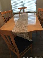 Photo of rectangular blonde wood dining table with leaf extension, four matching chairs, and a light-colored woven table runner laid lengthwise on the table. The table surface is glossy with visible scratches.