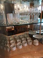 Porcelain dinnerware set displayed on middle and lower shelves including cups, saucers, plates, and a sugar bowl all monogrammed with gold trim.