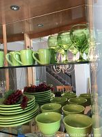 Green ceramic mugs and green glass goblet-style glasses displayed on a glass shelf inside a cabinet, reflections show dining room surroundings.