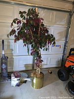 Artificial ficus tree in a brass planter with visible broken branch and a mix of green and reddish leaves. Shows some dust and dirt.