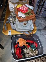 Overview of handbags stacked on a small side table and assorted fanny packs and wallets inside a gray plastic bin on the floor.