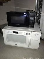 Photo showing the stacked small black Proctor Silex microwave on top of the large white Kenmore microwave with integrated toaster oven, visible control panels and doors.
