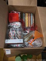 Box containing various plastic dinnerware items including square plates with a multicolor zigzag pattern, orange square bowls, and clear and orange tumblers, some still packaged.