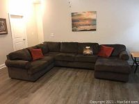Full view of L-shaped brown cloth sectional sofa with three pieces and several pillows, placed in living room space with wood flooring.