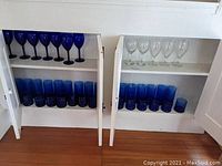 Two shelves of mainly cobalt blue and clear glassware inside a cabinet with doors open.