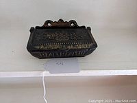 Front view of antique black cast iron wall mounted match safe with embossed floral designs and 'BARDAC' lettering on the lid.