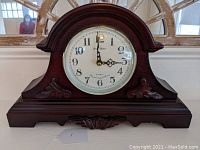Front view of the wooden mantle clock showing the curved top, carved decoration, white clock face with black numerals and clock hands with glass front.