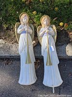 Two white blow mold angel Christmas lawn ornaments with gold wings and horns standing upright on grass.