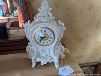 Front view of the ornate white porcelain clock on a wooden surface, showing full shape and sculpted details.