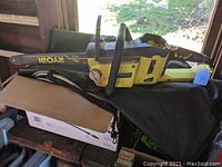 Ryobi 40V cordless electric chainsaw lying on a cloth covering a box beside a window, showing full side profile of tool including chain and bar, yellow and black colors visible.