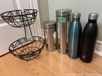Photo showing black metal two-tier basket with ornamental scroll design and four cylindrical items: two stainless steel canisters with lids, and two Manna insulated water bottles in blue and black colors.