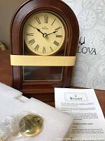 Wooden Bulova mantel clock with arched top, Roman numeral clock face, black hands, packaging foam, and instruction sheet visible.