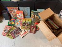 Overview of the assortment of children's books spread on the floor and partially packed in a cardboard box, showing a variety of titles and conditions.