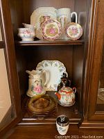 Wide shot of wooden cabinet shelf displaying assorted china, glass plates, and ceramic pieces including Royal Worcester egg coddler and Royal Albert cup and saucer