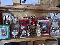 Shelf display showing various Christmas decorations including snowmen figurines, an angel, candle holders, a boxed hurricane candleholder set, and snow globes arranged on wood shelving.