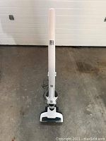 Upright view of the white and black Eureka Lightspeed cordless vacuum cleaner with its charging base and power cord on the ground behind it.