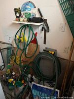 Photo showing wall mounted shelf with pruners, hand plant trowel, sprinkler parts, garden hose coiled and hanging, garden tools leaning on wall