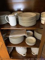 Shelves of fine china plates, cups, and serving pieces, showing stacked plates and cream jug inside a wooden cabinet.