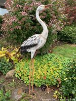 Side view of the metal crane yard ornament positioned in a garden bed with bushes and stone accents, showing the detailed feathers and coloration on the body and head.