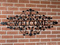 Full view of black metal welcome sign mounted on brick wall with intricate floral and leaf patterns.