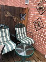 Two green metal patio chairs with green and white striped cushions, glass top round table, wooden fence and brick wall background with metal dragonfly and butterfly wall decorations