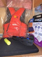 Photo showing orange BuoyOBuoy life vest, black seat pad, small yellow cylindrical object and boxed Kabooti 3-in-1 seat cushion