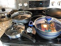 Group of pots and pans displayed on stove including Henckel poached egg pan, FlavorStone pot with lid, gray skillet with lid, and large wok.