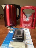 Red Hamilton Beach cordless electric kettle, bagel slicer, and packaged wireless thermometer displayed together on table.