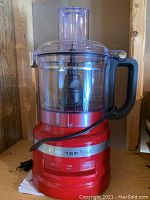 Front view of red KitchenAid food processor showing clear plastic bowl with handle, red base, chrome band with KitchenAid logo, and controls.