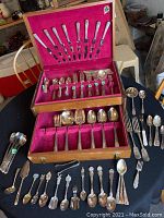 Full set of silver plate cutlery displayed in wooden box with red velvet lining, plus additional serving pieces and souvenir spoons spread on table.