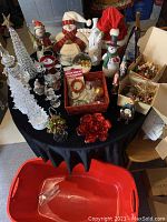 Photo showing table with large illuminated clear acrylic trees, assorted snowman figures, ornaments in boxes, and various holiday decorations spread on a black cloth covered table.