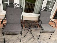 Photo of two black metal patio chairs and a small matching table on a tiled floor, showing grey seat cushions on chairs and decorative scroll design on chair backs.