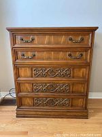 Front view of the tall boy dresser showing the wooden finish, four drawers with metal handles, and ornate carvings on three lower drawers.
