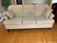 Front view of the beige upholstered sofa with tufted back and two throw pillows on a hardwood floor.