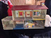 Front view of the vintage metal dollhouse with interior rooms visible, showing wallpapered rooms, a balcony, and a bag of dollhouse furniture inside the bottom right room.