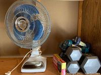 Photo showing white oscillating fan with blue blades, a variety of dumbbells including vinyl-coated and metal hexagonal weights, and stopwatch box on wooden shelf.