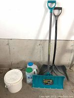 Two snow shovels, one blue plastic with metal shaft and one black plastic, each with different handle styles, standing against a wall with three containers at their base including a white bucket and two ice melter bottles.
