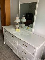 White painted dresser with nine drawers and brass handles topped with lace doily, two vintage white glass oil lamps with floral decoration and rectangular mirror with rounded corners.