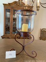 Photo shows a large floor candle holder with a 20 inch height. The candle holder has a curved metal base with ornamental scroll designs and a clear glass hurricane lamp containing a yellow pillar candle.