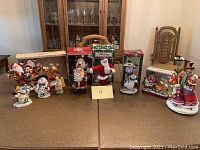 Nine Christmas figurines displayed on wooden tables. Includes multiple Santas, snowmen, and a musical Santa in boxed sleigh.