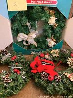 Full view of six Christmas wreaths with various decorations including red bows, pinecones, berries, snowflakes, and cream flowers. One wreath is boxed.