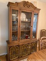 Large wooden dining hutch with ornate top, glass front upper cabinets with decorative metal lattice, and wood paneled lower cabinets with hardware handles. Shelves inside are clear glass, containing visible glassware and dishes (not included).