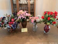 Photo showing four artificial floral arrangements on a table, including large basket and wood-look urn, plus two clear glass vases.