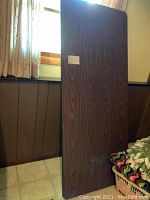 Front view of rectangular dark brown folding table top leaning upright against a wall with wood paneling and window above.