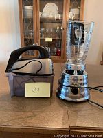 Photo showing the hand mixer in black and silver with its storage container next to a chrome blender base with glass pitcher and black lid, placed on a wooden table in front of a china cabinet.