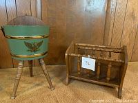 Two magazine racks side by side; one green painted bucket style with gold eagle emblem on tripod legs, and one brown spindle wooden rack.